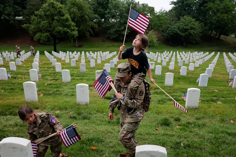 A member of the 3rd U.S. Infantry Regiment with her daughter on her shoulders places flags in front of each headstone at Arlington National Cemetery on May 21, 2026 in Arlington, Virginia. Before dawn soldiers begin the process of placing a flag in front of approximately 260,000 headstones ahead of Memorial Day.